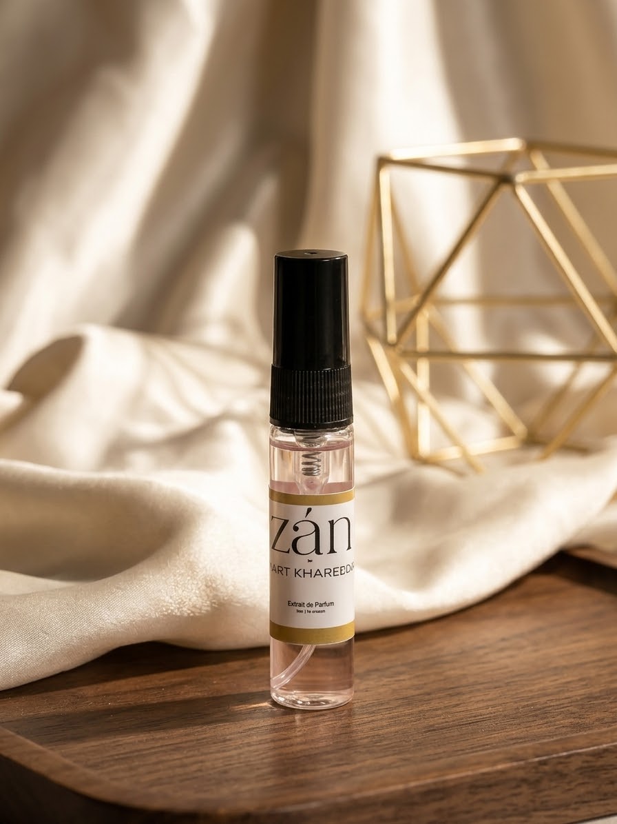 A small clear spray bottle with a black cap stands on a wooden surface, surrounded by draped cream fabric and a gold geometric decor object in the background. The label reads 'zán' and 'MAINT-KHAREEBA'.